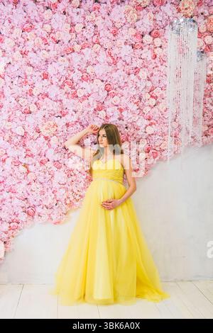 Eine junge kaukasische Frau mit langen braunen Haaren posiert in einem gelben Kleid vor einem Hintergrund rosafarbener Blumen. Sie hält ihren Bauch und schlägt eine Mutterschaft vor Stockfoto