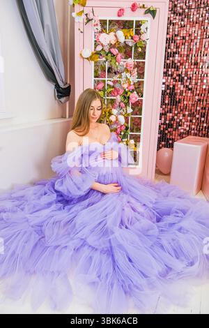 Eine junge kaukasische Frau mit langen braunen Haaren sitzt anmutig in einem Kleid aus Lavendel-Tüll. Sie ist in einem dekorierten Zimmer mit Blumenakzenten und rosa Rückseite Stockfoto