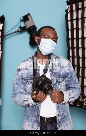 Porträt eines schwarzen Kameramanns, der sich mit einer Gesichtsmaske schützt, während er im Studio mit blauem Hintergrund steht. afroamerikanischer Fotograf, der Nase und Mund bedeckt und seine dslr-Kamera hält. Stockfoto