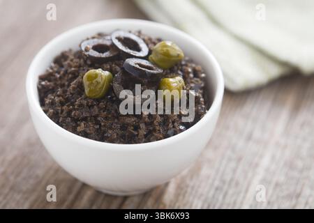 Hausgemachter französischer Tapenade-Aufstrich aus schwarzem Oliven, garniert mit Olivenscheiben und Kapern, serviert in einer kleinen Schüssel (selektiver Fokus) Stockfoto
