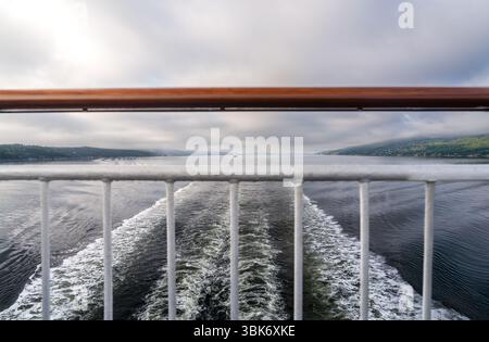 Morgens bricht warmes Licht durch den Nebel über Oslofjord, Norwegen. Ich schaue durch das Geländer auf dem Hinterdeck mit einem Kreuzfahrtschiff in seiner Spur Stockfoto