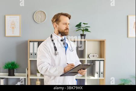 Porträt eines jungen selbstbewussten männlichen Arztes in weißer medizinischer Uniform, der in seiner Praxis steht Stockfoto