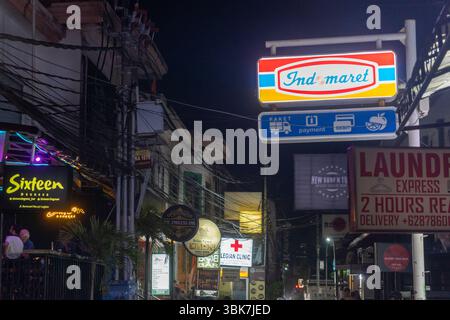 Bali, Indonesien - 25. Dezember 2024: Mann sitzt auf dem Bürgersteig vor dem Indomaret-Supermarkt. Die Straße ist dunkel. Stockfoto