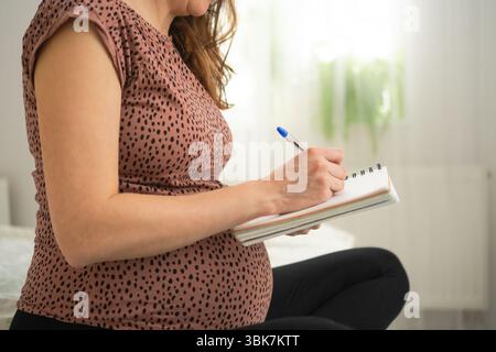 Eine schwangere Frau schreibt in ein Notizbuch, während sie auf dem Bett sitzt. Planung von Baby-Essentials und Krankenhaustasche. Stockfoto