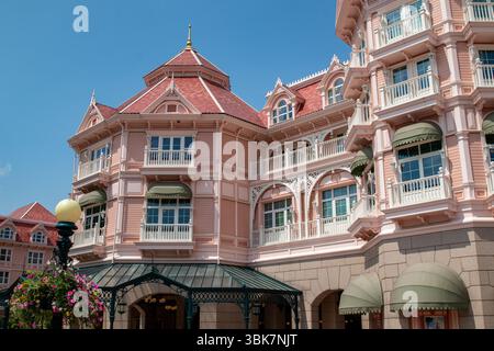 Fassade eines rosafarbenen Gebäudes mit weißer Zierleiste, rotem Ziegeldach, grünen Markisen und Balkonen unter klarem blauen Himmel im Disneyland Paris. Stockfoto