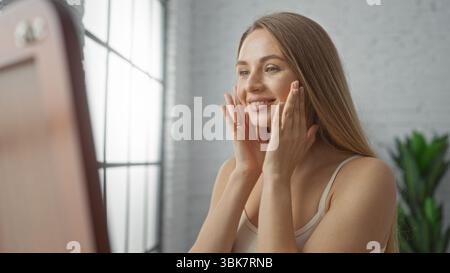 Frau, die Reflexion im hellen Wohnzimmer bewundert, mit glatter, gesunder Haut, mit natürlichem Licht, das durch große Fenster strömt und Schönheit verkörpert Stockfoto