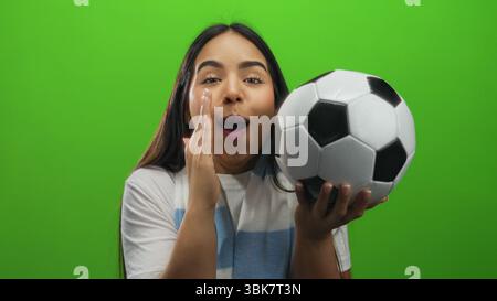 Aufgeregte junge Frau, die einen Fußball hält und drinnen vor einem lebendigen grünen Hintergrund mit ausdrucksstarkem Lächeln feiert, das Freude und hauch vermittelt Stockfoto