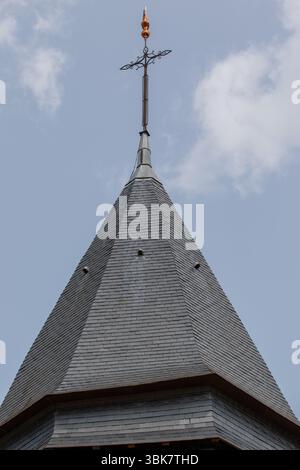 Bild eines hohen, steilen, grauen Schieferdachs, gekrönt mit einem Metallkreuz und dekorativem Finial, vor einem blauen Himmel mit verstreuten Wolken in Giverny Stockfoto