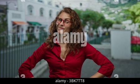 Selbstbewusste Frau in rotem Hemd steht mit Armen akimbo auf einer Stadtstraße und vermittelt Entschlossenheit und Selbstbewusstsein in einer urbanen Outdoor-Umgebung. Stockfoto