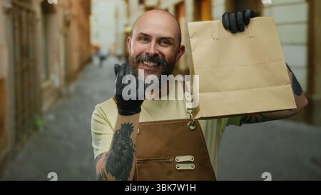 Ein lächelnder Mann hält Papiertüte auf der städtischen Straße mit Schürztätowierungen und Handschuhen, was auf einen modernen Lieferservice im Freien hindeutet. Stockfoto