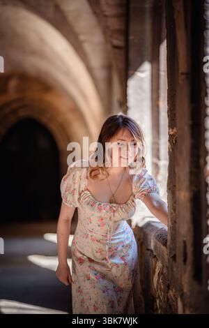 Eine junge Frau in einem Blumenkleid posiert anmutig in der Nähe eines mittelalterlichen Steinbogens mit sanfter Innenbeleuchtung. Stockfoto