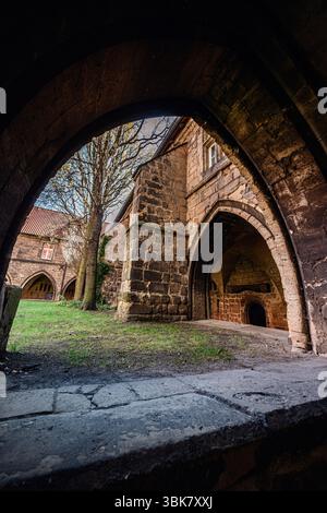 Der Blick durch einen dunklen Steinbogen offenbart einen grasbewachsenen Innenhof, der von einer bogenförmigen mittelalterlichen Architektur und mit Efeu bedeckten Wänden umgeben ist. Stockfoto