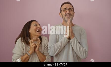 hispanische Paare mittleren Alters, die Liebe und Humor mit gekreuzten Daumen und Händen an einer pinkfarbenen Wand ausdrücken. Stockfoto