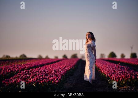Eine Frau in einem Blumenkleid steht friedlich in einem blühenden Tulpenfeld und sonnt sich im warmen Glanz des Sonnenuntergangs. Stockfoto
