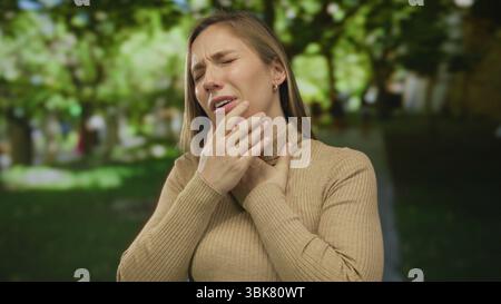 Frau draußen im Park, die Beschwerden oder Schmerzen zeigt, Kehle umklammert, umgeben von Bäumen und Gras an einem sonnigen Tag in lässiger Kleidung. Stockfoto