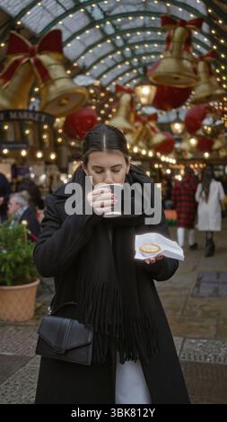 Frau genießt festliche Atmosphäre im london covent Garden mit weihnachtslichtern, die Kaffee und Gebäck unter verzierten Bögen halten. Stockfoto