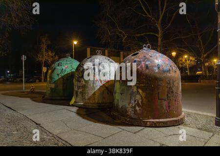 Drei mit Graffiti bedeckte Recycling-Abfallbehälter aus Glas säumen nachts einen Bürgersteig, beleuchtet von Straßenlaternen in einem urbanen Ambiente. Berlin, Deutschland. 29. März 2025. Stockfoto