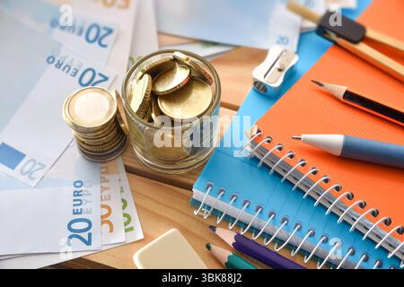 Detail der Ausgaben für Schulmaterialien mit Geld, Notizbüchern und Farbstiften auf einem Holztisch. Erhöhte Aussicht. Stockfoto