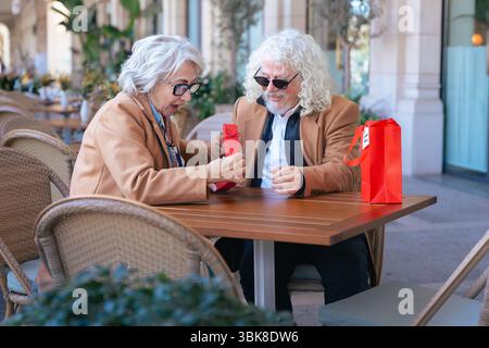 Lächelndes Seniorenpaar genießt gemeinsam Kaffee im Café im Freien in lässigen Outfits Stockfoto