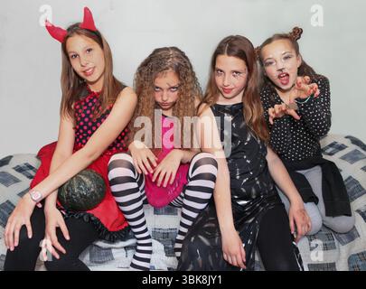 Vier junge Mädchen in farbenfrohen Halloween-Kostümen posieren drinnen, lächeln und blicken in die Kamera. Stockfoto