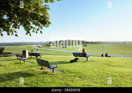 Menschen sitzen auf Bänken in der Sonne auf dem Hügel in Tattenham Corner mit Blick auf die berühmte Rennbahn Epsom Downs, Heimstadion des Derby, Surrey England, Großbritannien Stockfoto