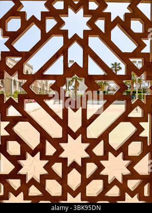 Nahaufnahme einer traditionellen arabischen Holzwand mit sternförmigen Ausschnitten, die einen hellen Innenhof mit Palmen im Hintergrund umrahmt. Toll für cultura Stockfoto