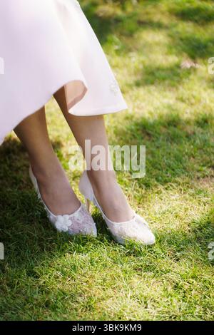 Nahaufnahme der Beinchen der Frau, die zarte Spitzen-High-Heels mit Blumendetail tragen und während der Zeremonie am Hochzeitstag auf grünem Gras im Sonnenlicht warten Stockfoto