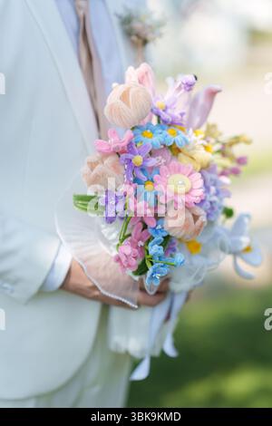 Großaufnahme des Bräutigams in weißem Anzug, der während der Hochzeitsfeier eine künstliche Blumenstraußblume in Pastellfarben hält Stockfoto