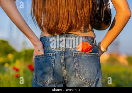 Frau in Jeans mit Poppy Flower draußen. Nahaufnahme einer jungen Frau, die an einem sonnigen Tag auf einem Feld mit einer roten Mohnblume in ihrer Jeanstasche steht. Stockfoto