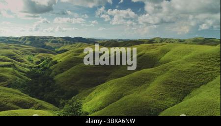 Aus der Vogelperspektive die hügelige Landschaft von Sumba Island, Indonesien, mit sanften grünen Hügeln, die sich so weit das Auge reicht, unter einem dramatischen Himmel mit flauschigen weißen Wolken Stockfoto
