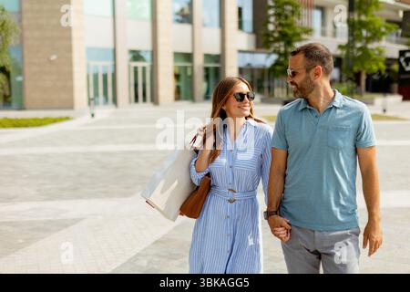 Ein Paar schlendert Hand in Hand durch ein zeitgenössisches Einkaufsviertel, sonnt sich in der warmen Sonne und teilt freudige Gespräche. Stockfoto