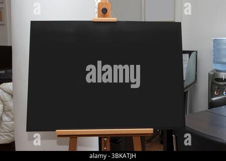 Leere schwarze Leinwand auf einer Holzstaffelei in einem modernen Bürozimmer mit Möbeln und Wasserspender, Konzept der Kreativität oder Präsentation Stockfoto