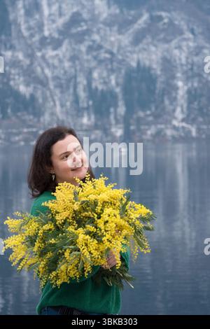 Eine Frau mit Mimosa-Blumen am Ritsa-See in Abchasien, die vor einem Hintergrund von Bergen und Wasser steht Stockfoto