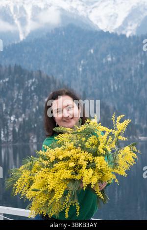 Eine Frau mit Mimosa-Blumen am Ritsa-See in Abchasien, die vor einem Hintergrund von Bergen und Wasser steht Stockfoto