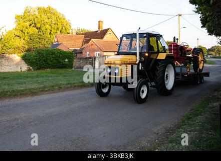 Leyland 802 Traktor schleppt eine antike rote Landwirtschaftsmaschine auf einer Landstraße mit traditionellen Backsteinhäusern im Hintergrund Stockfoto