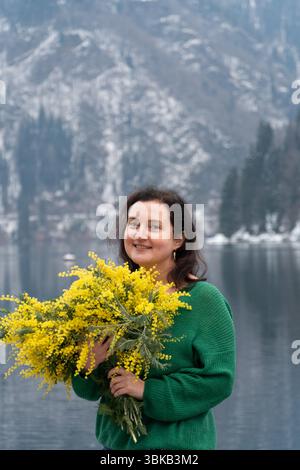 Eine Frau mit Mimosa-Blumen am Ritsa-See in Abchasien, die vor einem Hintergrund von Bergen und Wasser steht Stockfoto