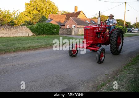 Ein Mann, der einen alten roten Traktor im Poulshot Wiltshire Oldtimer fährt, trifft sich bei Sonnenuntergang mit traditionellen Backsteinhäusern im Hintergrund Stockfoto