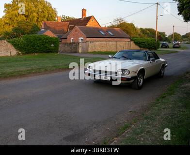 Der Jaguar XJS Sportwagen in Oldtimer-Creme fährt bei Sonnenuntergang an bezaubernden englischen Landhäusern vorbei Stockfoto