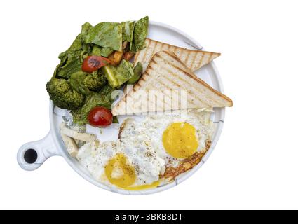 Zwei gebratene Eier mit gedünstetem Gemüse und Toast Brot auf einer weißen Keramikpfanne, Blick von oben, isoliert Stockfoto
