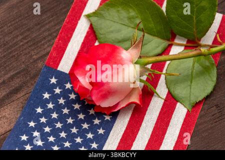 Nahaufnahme einer roten Rose, die auf der Flagge der Vereinigten Staaten von Amerika ruht, vor dunklem Holzhintergrund. Das Bild symbolisiert Erinnerung, Re Stockfoto