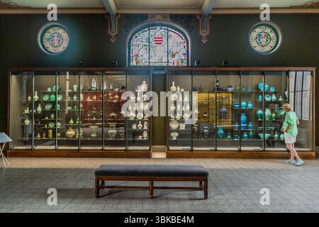 Eine Vitrine mit nach Farbe sortierten Porzellanobjekten steht im Musée National Adrien Dubouché am Place Winston Churchill in Limoges, Haute-Vienne, Nouvelle-Aquitaine, Frankreich. Zwei lange Glasschränke enthalten Vasen, Krüge, Teller und Figuren, die von weiß und celadongrün links zu dunklen, türkisfarbenen und blauen Glasuren rechts angeordnet sind. Die Journalistin Angela Berg, die einen Strohhut, eine hellgrüne Bluse und weiße Shorts trägt, steht am rechten Schrank und betrachtet die Objekte. Über den Schränken befinden sich ein gewölbtes Buntglasfenster mit einem Wappen und zwei runden Buntglasmedaillons. Stockfoto
