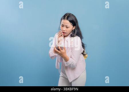 Eine Frau schaut auf ihr Handy, während sie vor einer blauen Wand steht. Sie ist von etwas auf ihrem Handy überrascht oder schockiert Stockfoto