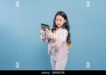 Eine Frau hält eine Tablette in der Hand und sieht sie mit einem überraschenden Ausdruck an. Sie trägt ein pinkfarbenes Hemd und eine weiße Hose Stockfoto