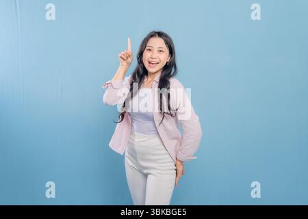 Eine Frau steht vor einer blauen Wand mit einem rosa Hemd und einer weißen Hose. Sie zeigt mit dem Finger nach oben und lächelt Stockfoto