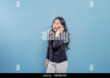 Eine Frau spricht auf ihrem Handy, während sie vor einer blauen Wand steht. Sie ist überrascht oder schockiert von etwas, das sie am Telefon hört Stockfoto