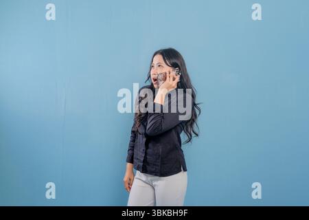 Eine Frau spricht auf ihrem Handy, während sie vor einer blauen Wand steht. Sie ist überrascht oder schockiert von etwas, das sie am Telefon hört Stockfoto