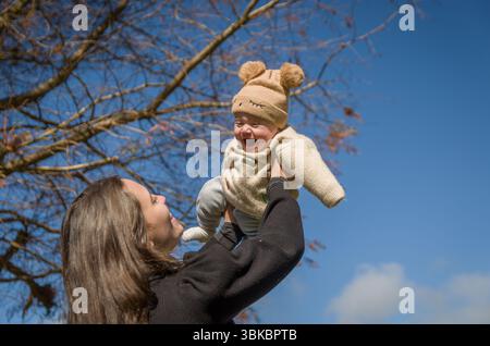 Mutter spielt mit dem Sohn auf dem sonnigen Platz und wirft ihn auf, während er lächelt. Stockfoto