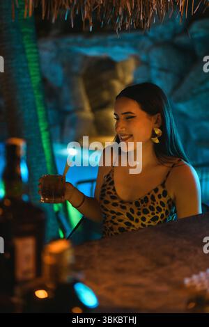 Stilvolle Brünette Frau im Leopardenkleid, die Getränke in der eleganten Hotelbar mit Pool im Hintergrund genießt. Stockfoto