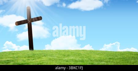 Flaches, verwittertes Holzkreuz steht auf einem abfallenden grünen Hügel unter blauem Himmel mit Sonnenstrahlen Stockfoto
