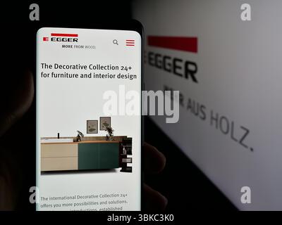In dieser Fotoabbildung hält eine Person ein Smartphone mit der Webseite des österreichischen Unternehmens Egger Holzwerkstoffe GmbH auf dem Bildschirm vor dem Logo. Stockfoto